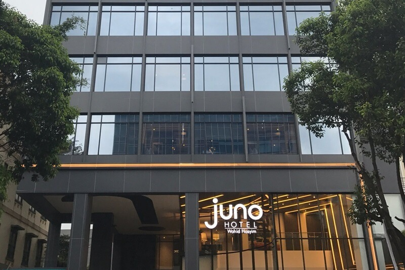 Juno Hotel entrance sign with modern glass office building facade and trees framing the storefront.