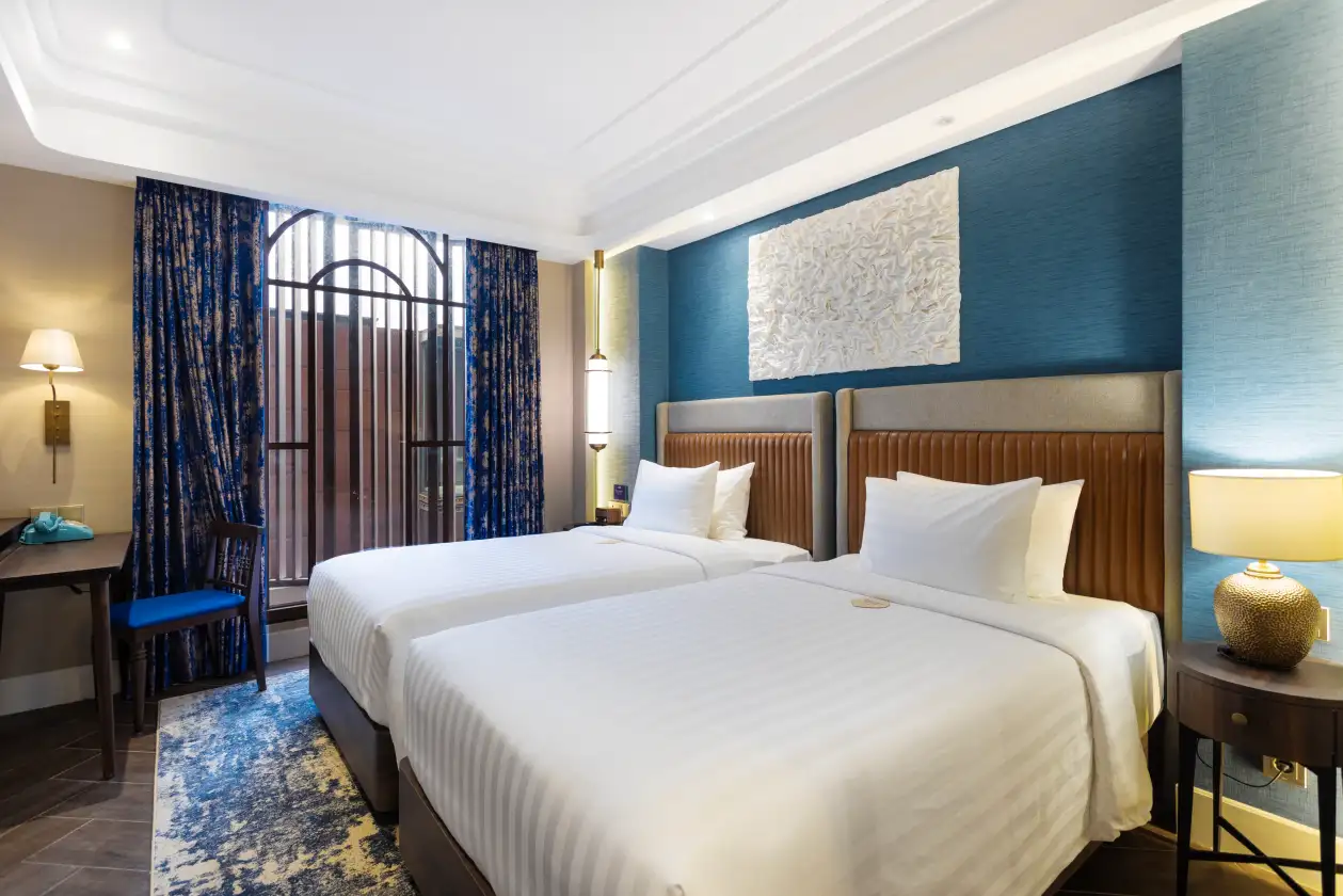 Modern hotel bedroom with twin beds, blue curtains, upholstered headboards, bedside lamps, and blue accent wall