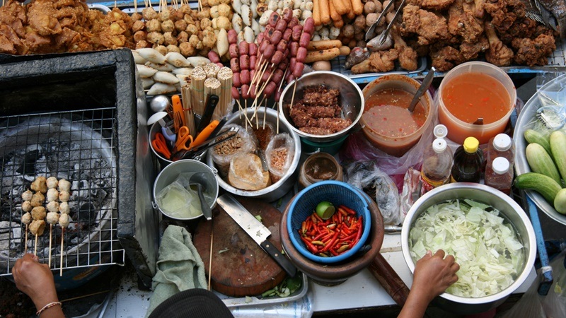 Street food stall with skewers, grilled meat, bowls of sauces and chopped vegetables on a crowded table