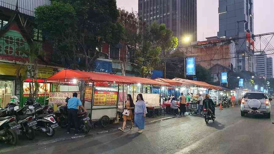 Street food stalls with red canopies and diners along a city sidewalk beside parked scooters and cars at dusk